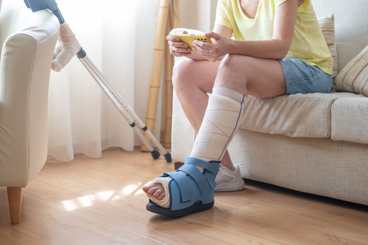 Mature woman sitting on a sofa, wearing an orthopedic boot to protect her injured and fractured foot.